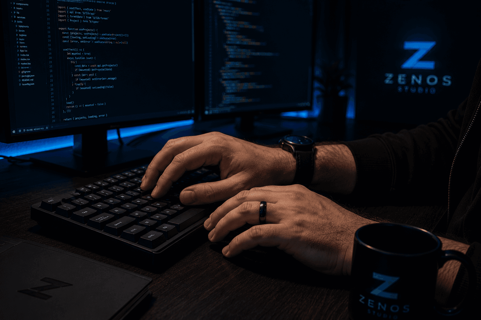 Mãos trabalhando no teclado com monitores de código e identidade da Zenos Studio ao fundo